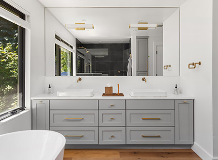 Double vessel sink vanity with gray cabinets next to a picture window and freestanding tub
