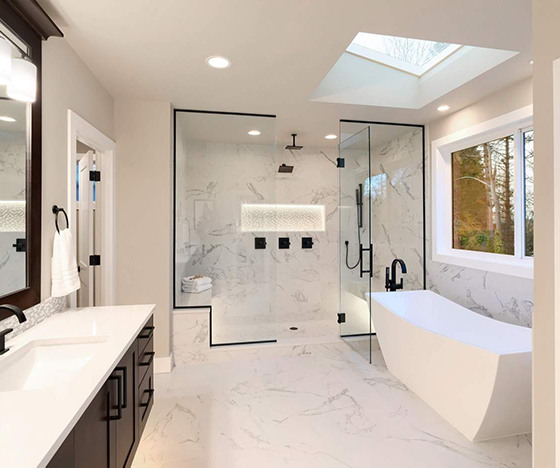 Large marble bathroom with large glass door shower and dark cabinet vanity and a freestanding tub beneath a skylight and next to a window