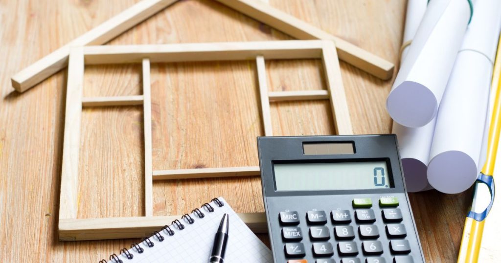 Small wooden house model with a calculator and notebook, representing construction budgeting or planning.