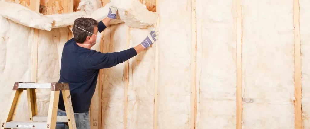 Worker installing fiberglass insulation in a wooden wall frame.