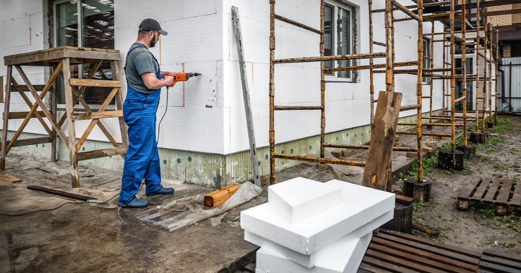 A construction worker in blue overalls and a baseball cap drilling into the white foam insulation of a home's exterior wall next to scaffolding during a major renovation
