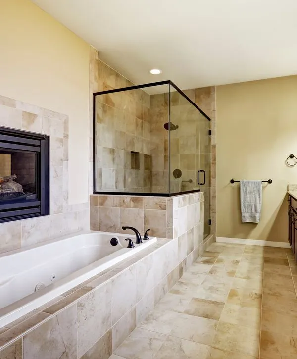 A luxurious primary bathroom featuring a large soaking tub next to a tiled fireplace, a glass-enclosed shower with beige tile, and matching neutral tile flooring for a cohesive look.