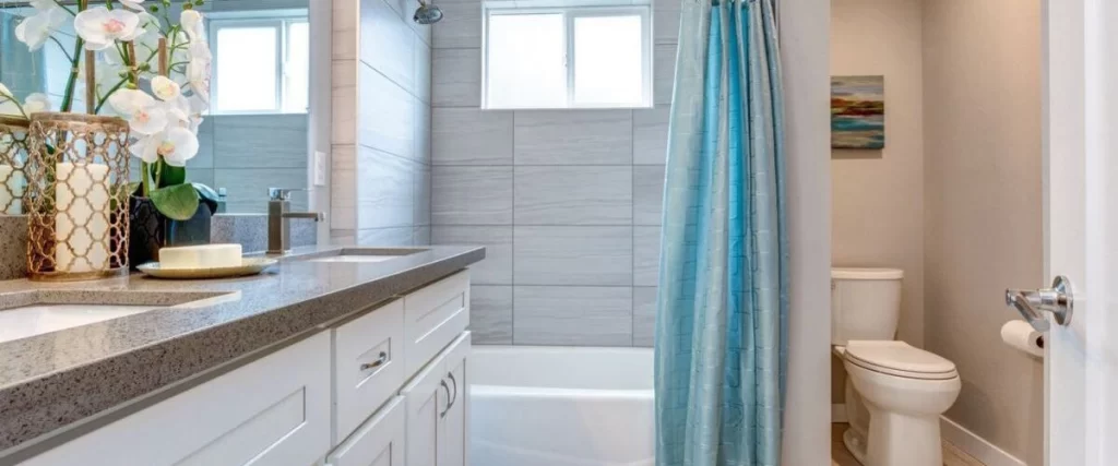 A functional small bathroom featuring a white vanity with a gray countertop and dual sinks, light gray tiled walls, and a blue shower curtain over the tub