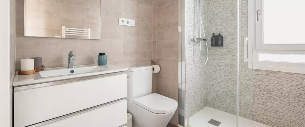 A clean contemporary bathroom showing a white double-drawer vanity next to a toilet and a walk-in shower with textured grey wall tiles and a rain shower head.