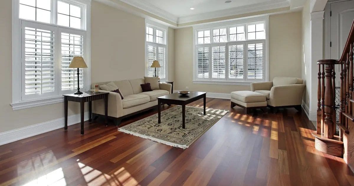 A spacious, sunlit living room featuring dark wood-look tiles, white plantation window shutters, a beige sofa, and a classic area rug.