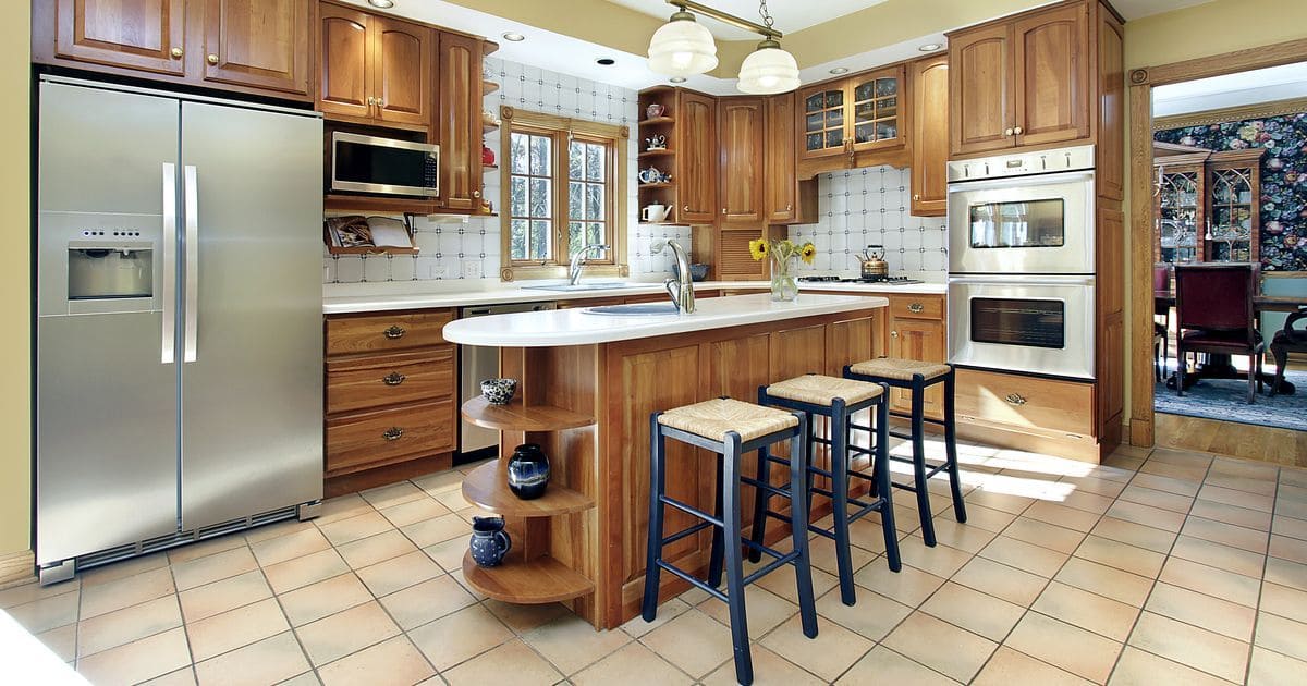 A spacious kitchen showcasing traditional kitchen trends with classic honey oak cabinets, a white central island, stainless steel appliances, and terracotta floor tiles.