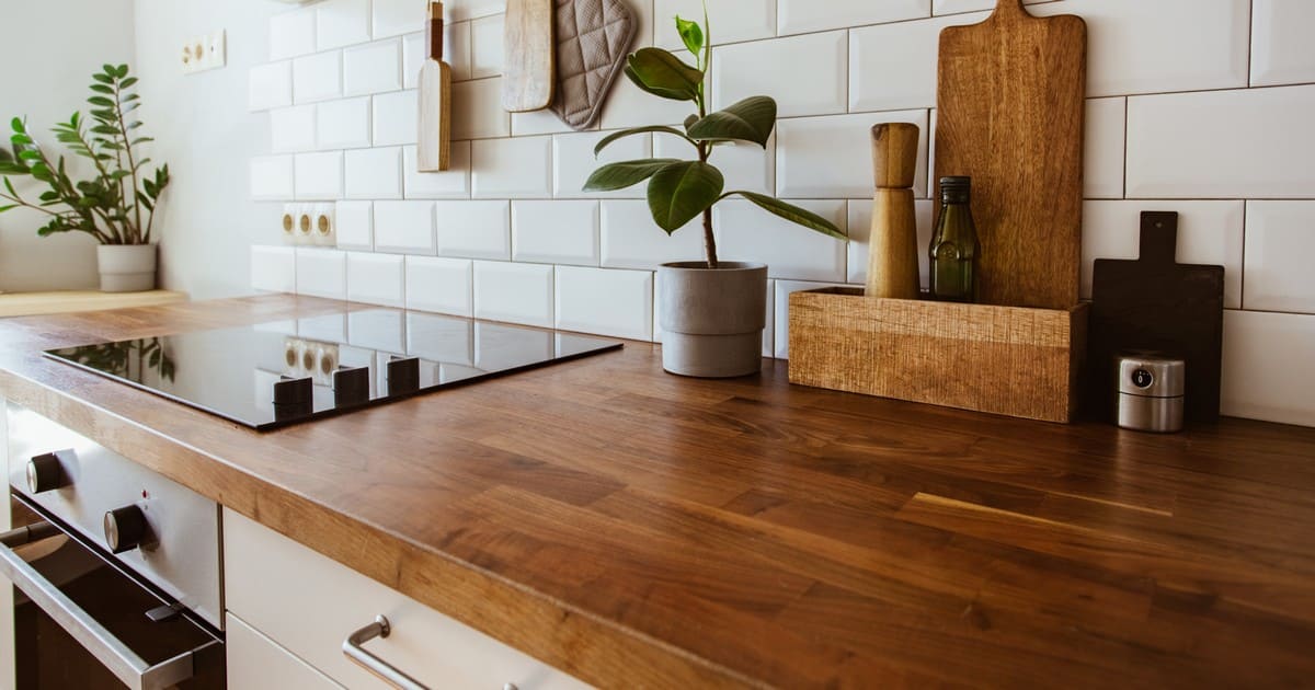 A close-up view of a warm natural wood kitchen countertop featuring a built-in stovetop, wooden cutting boards, and a small potted plant against a white tiled wall.