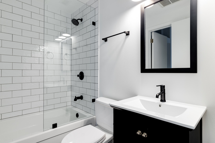 A-small-modern-bathroom-with-a-dark-vanity-mirror-frame-and-hardware.-White-subway-tiles-line-the-bathtub-and-shower-with-black-faucets