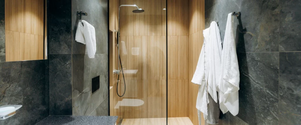 A modern walk-in shower featuring vertical wood-look porcelain tile contrasting with dark grey wall tiles and hanging white bathrobes.