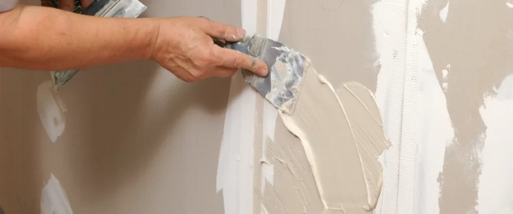 Close-up view of a worker's hands using a metal putty knife to smooth wet joint compound over fiberglass mesh tape on a panel of mold-resistant drywall.