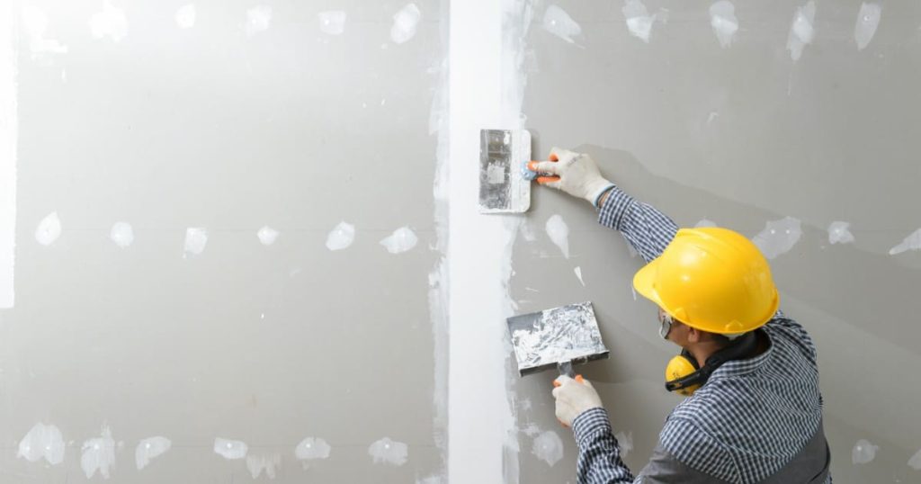 A construction worker wearing a yellow hard hat and safety glasses applying joint compound to a wall seam, illustrating Why West Phoenix Homes Need Mold-Resistant Drywall to protect against moisture.