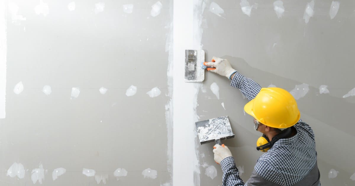 A construction worker wearing a yellow hard hat and safety glasses applying joint compound to a wall seam, illustrating Why West Phoenix Homes Need Mold-Resistant Drywall to protect against moisture.